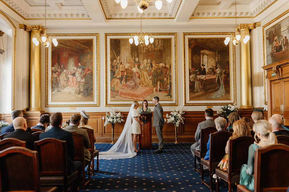 Bride and groom exchange vows inside Edinburgh City Chambers registry office surrounded by guests, beneath ornate gold-framed murals.