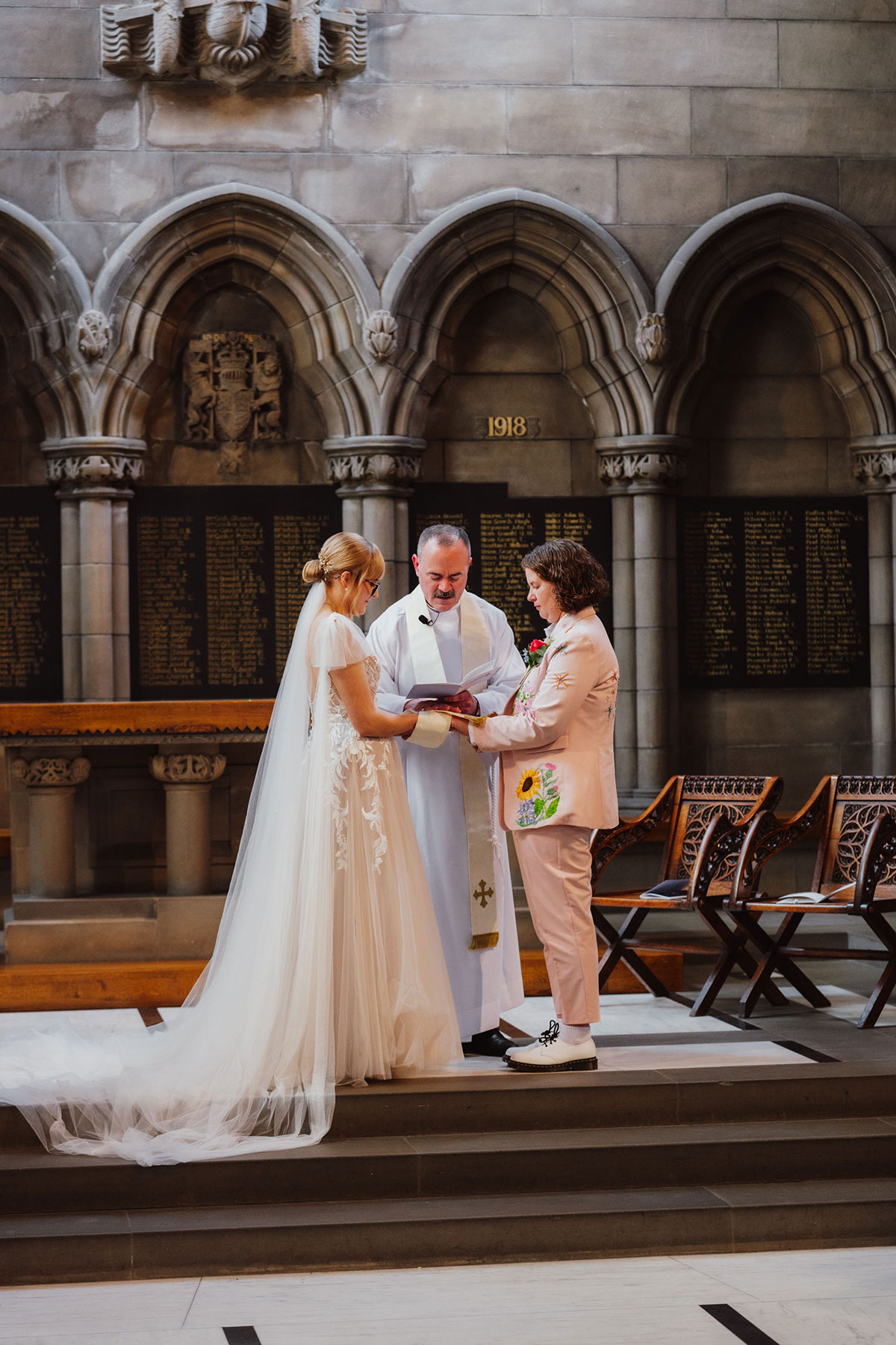 Couple during a ceremony inside the University of Glasgow Memorial Chapel, standing with the priest beneath arched stonework.
