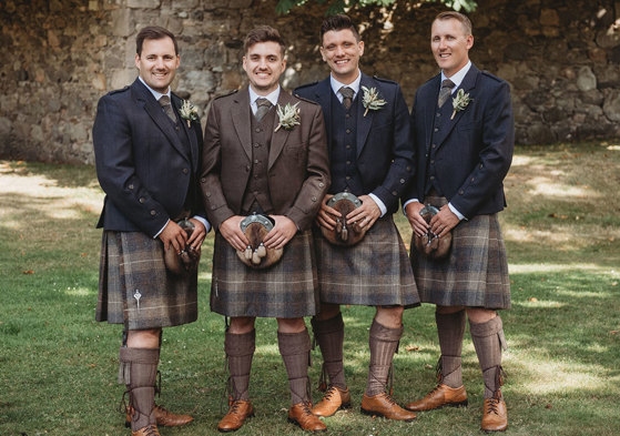 Four men standing outside wearing matching brown kilts, three with navy jackets and one with a brown jacket