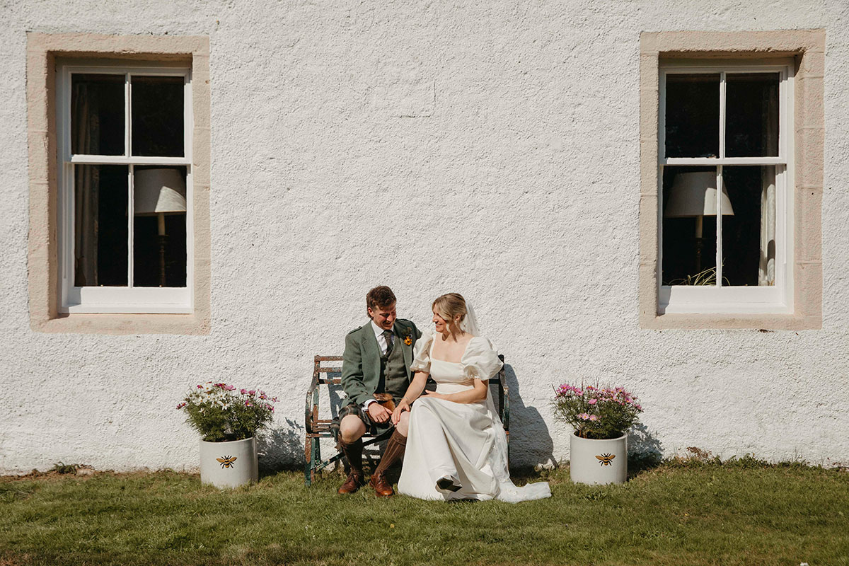 Bride and groom sit together on wooden bench outside Highland farmhouse wedding venue in Nethy Bridge
