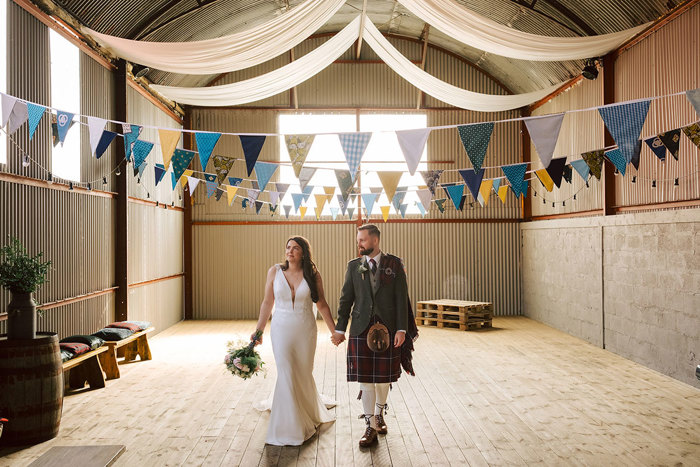 A bride holding a bouquet and a groom walk hand in hand through a large barn 