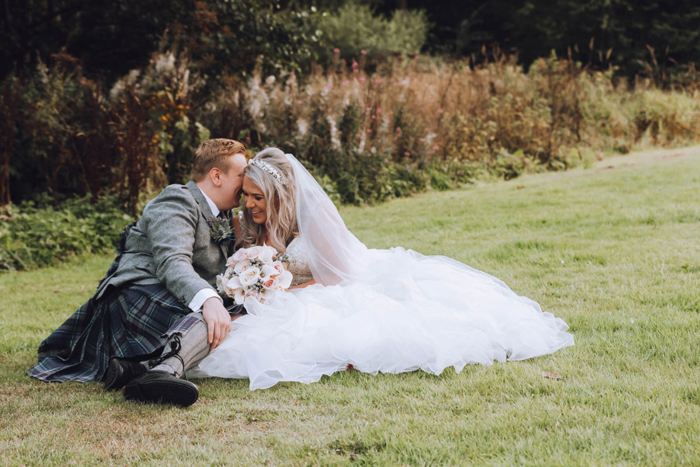 A Bride And Groom Sitting On Grass Whispering