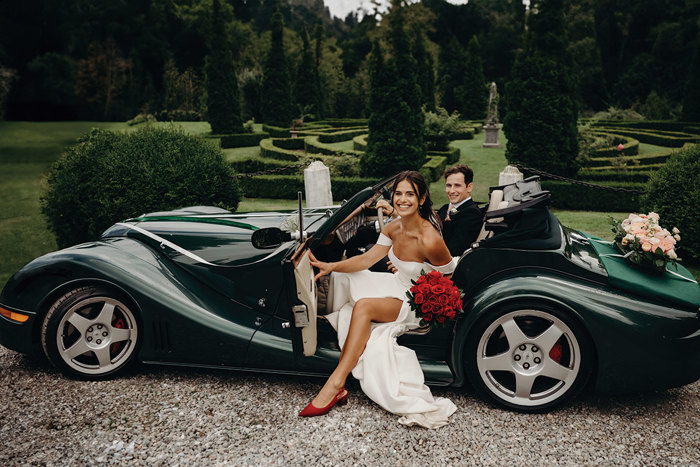 a bride stepping out of an open-top racing green sports car with the groom in the driving seat behind her. She is carrying a bouquet of red roses. The gardens of Achnagairn Castle are in the background.