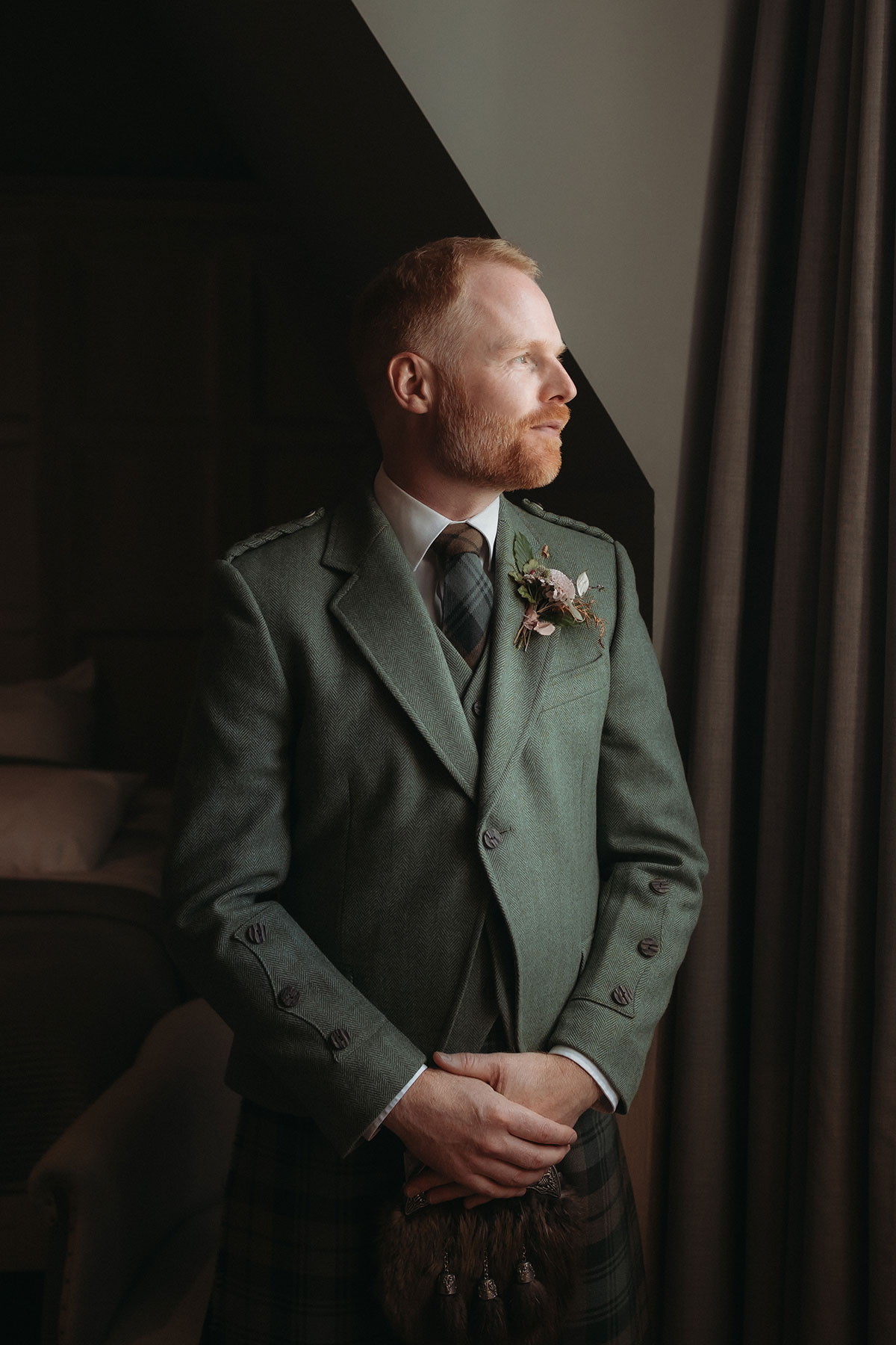 a man wearing a green tweed kilt jacket and tartan tie looking out of a window.