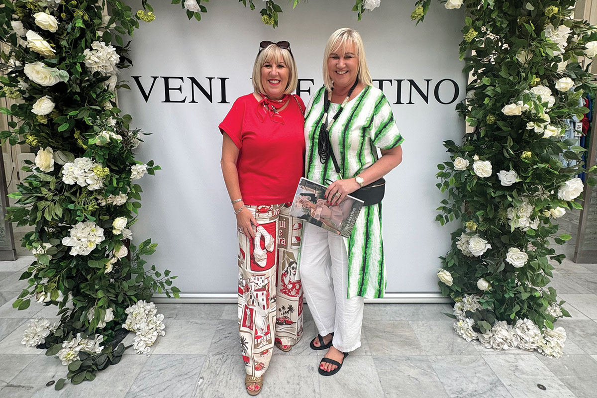 Two women standing in front of a floral backdrop with Veni Infantino branding, smiling at the camera inside a boutique setting