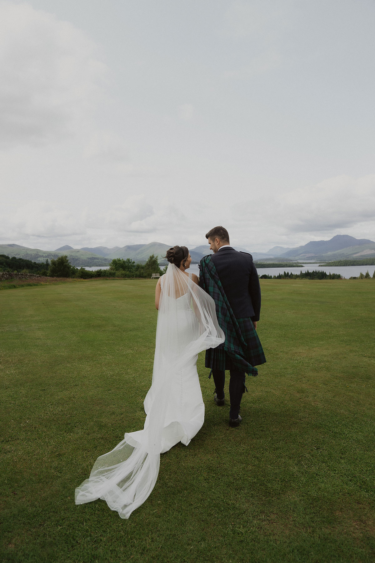 Bride and groom walking across Scottish countryside with sweeping loch and Highland mountain views behind them