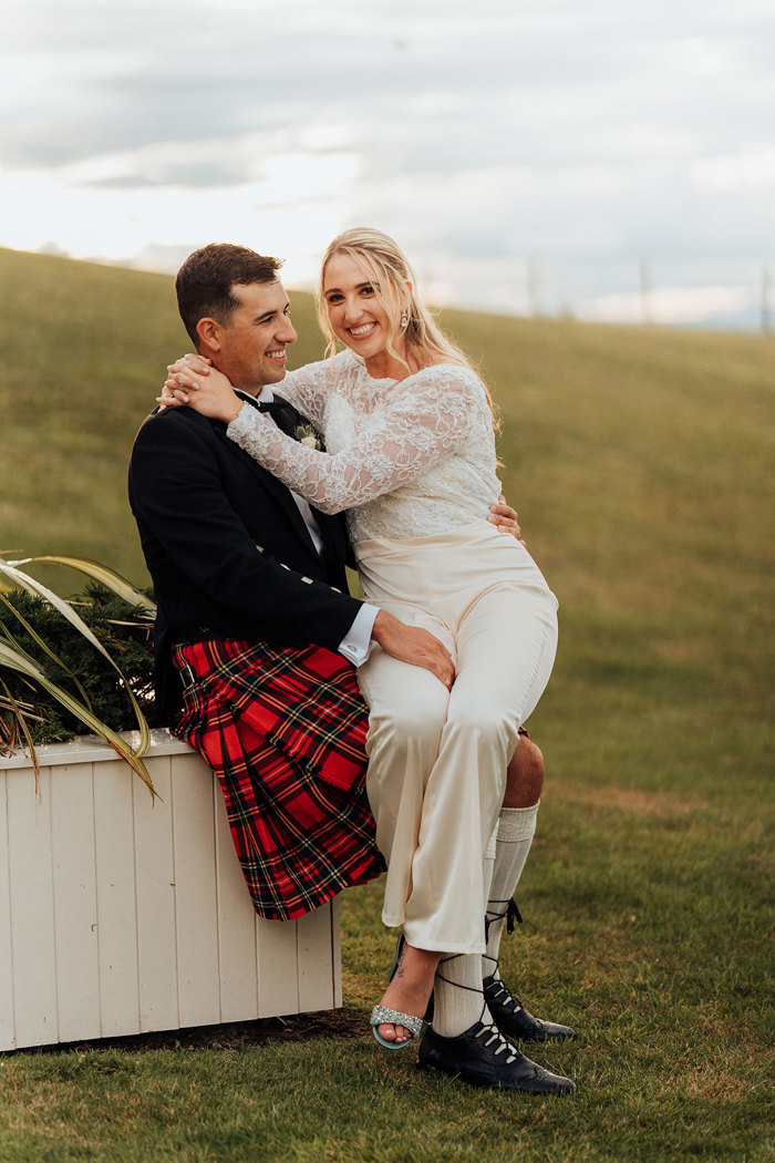 A bride wearing trousers and a groom wearing a red kilt sitting on a white fence.