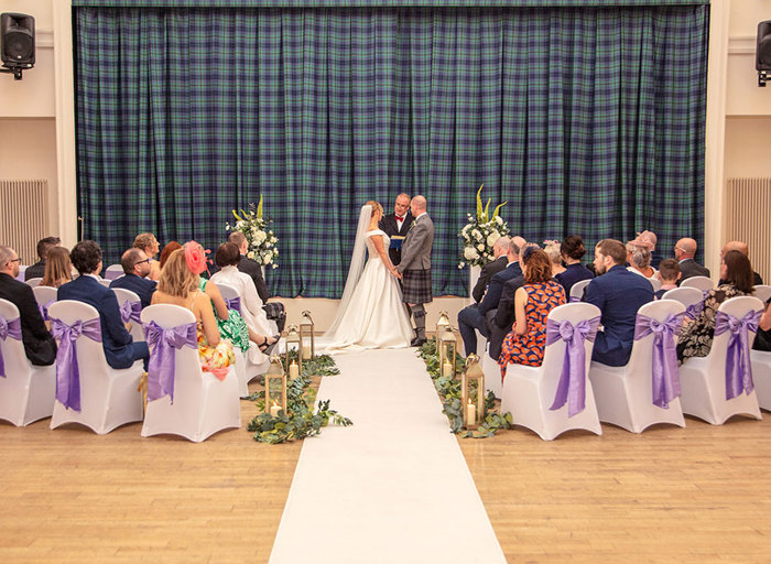 wedding ceremony with a green and blue tartan curtain backdrop, white chairs with purple bows, and a bride and groom and officiant standing at the end of the aisle