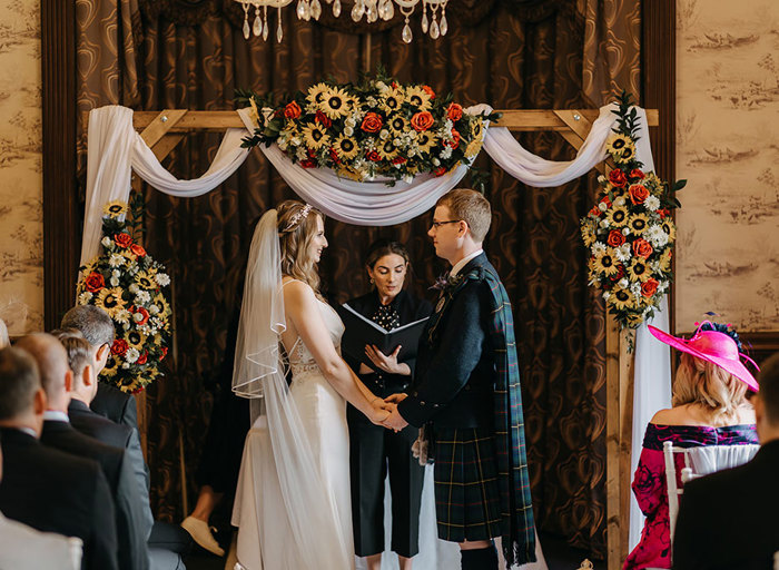 A wedding ceremony with a bride and groom holding hands standing in front of a celebrant who is reading from a book and standing underneath a wooden arch