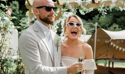 an elated bride and groom pop a champagne bottle in a garden setting with canvas stretch tent, wooden structure and greenery in background