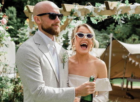 an elated bride and groom pop a champagne bottle in a garden setting with canvas stretch tent, wooden structure and greenery in background