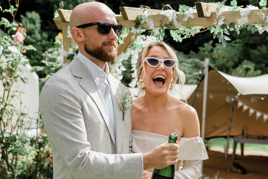 an elated bride and groom pop a champagne bottle in a garden setting with canvas stretch tent, wooden structure and greenery in background