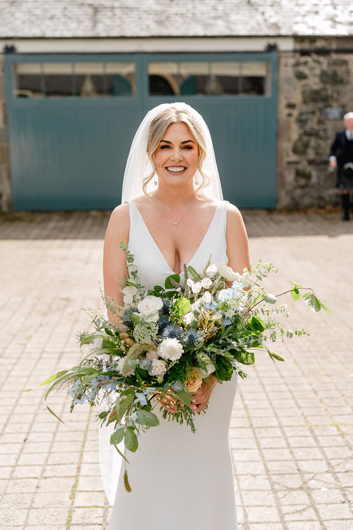 blonde smiling bride wears long veil in her hair, v-neck wedding dress and holds a wildflower bridal bouquet curated with lots of green, blue and thistles