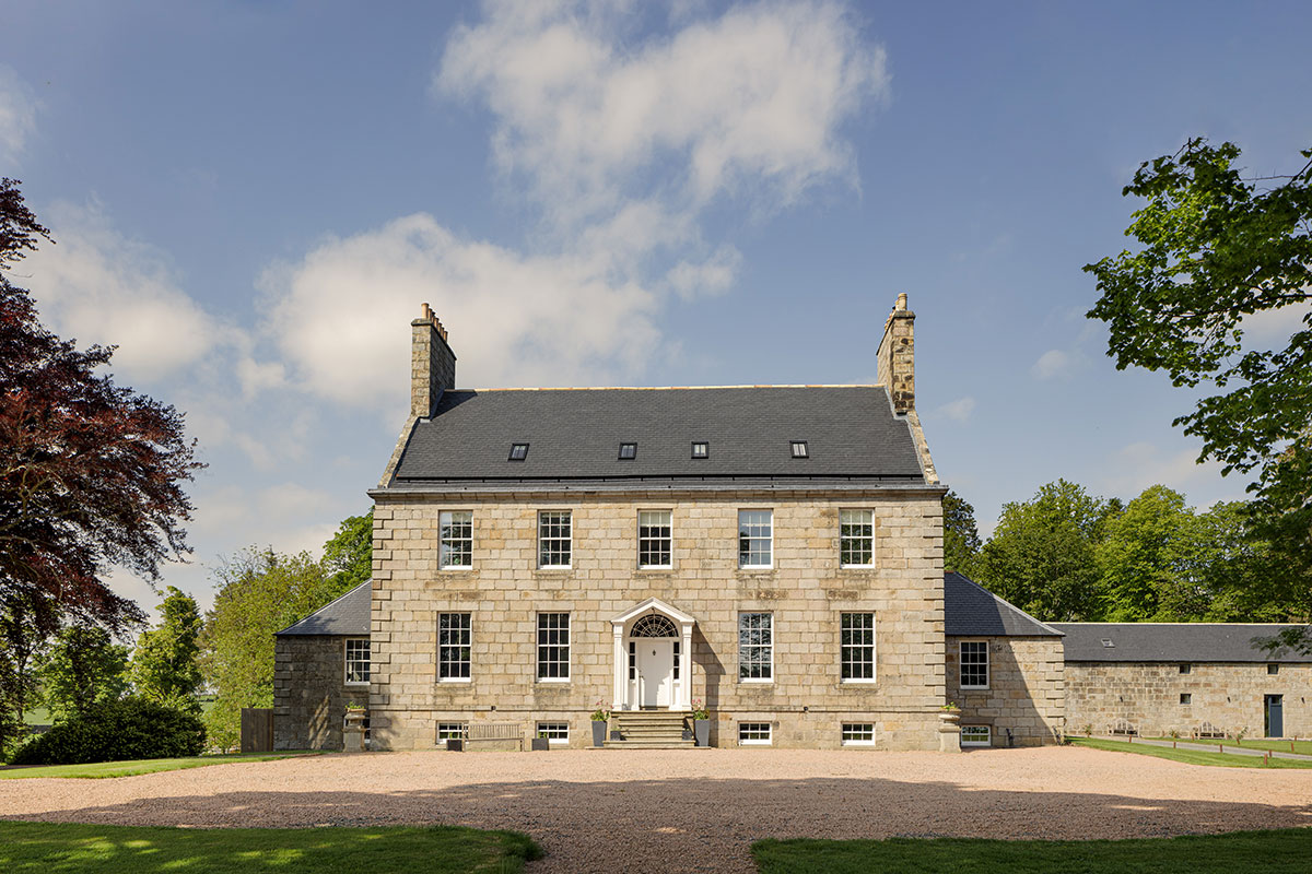 Front exterior of a large stone Georgian-style house with symmetrical windows, gravel driveway, and trees on either side
