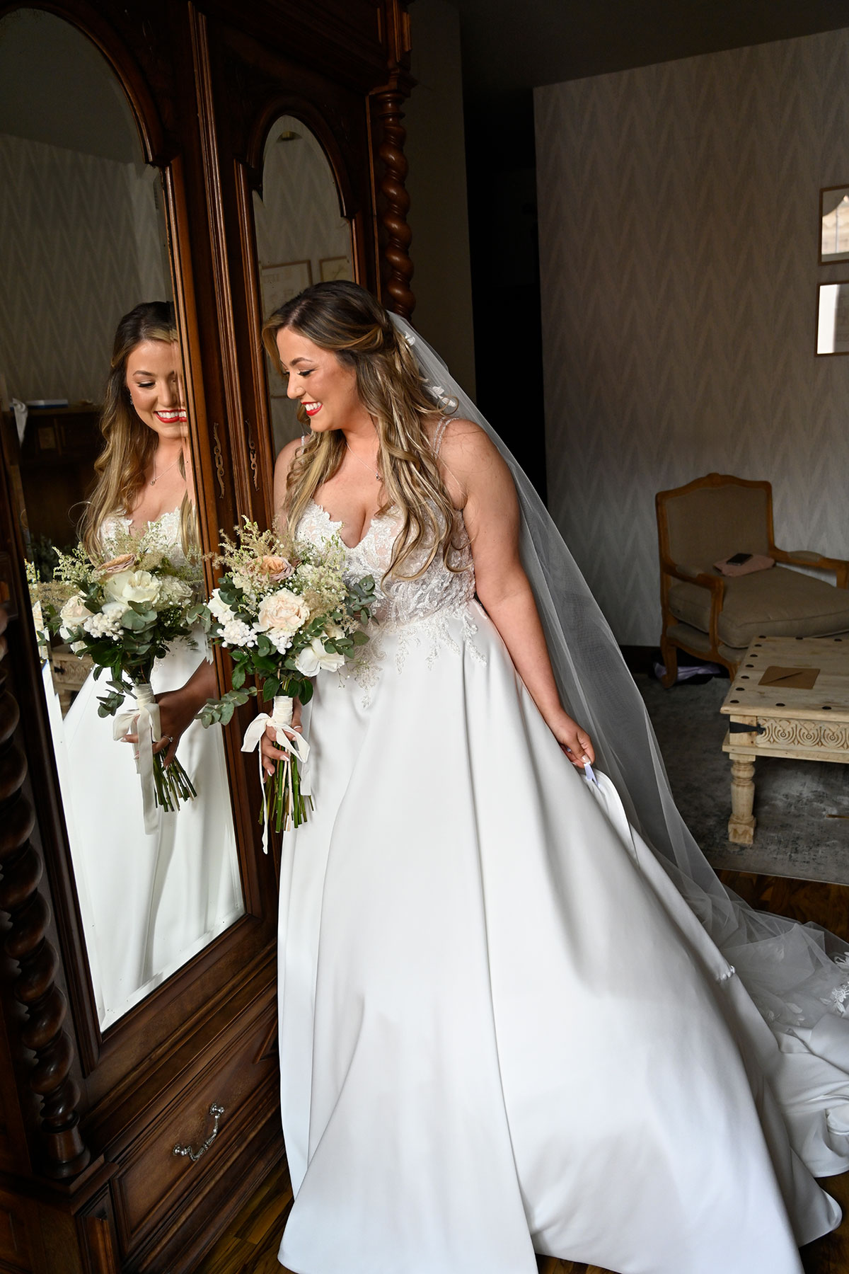 bride in ballgown wedding dress and trailing veil holds white bouquet and the back of her dress as she looks into wardrobe mirror at reflection