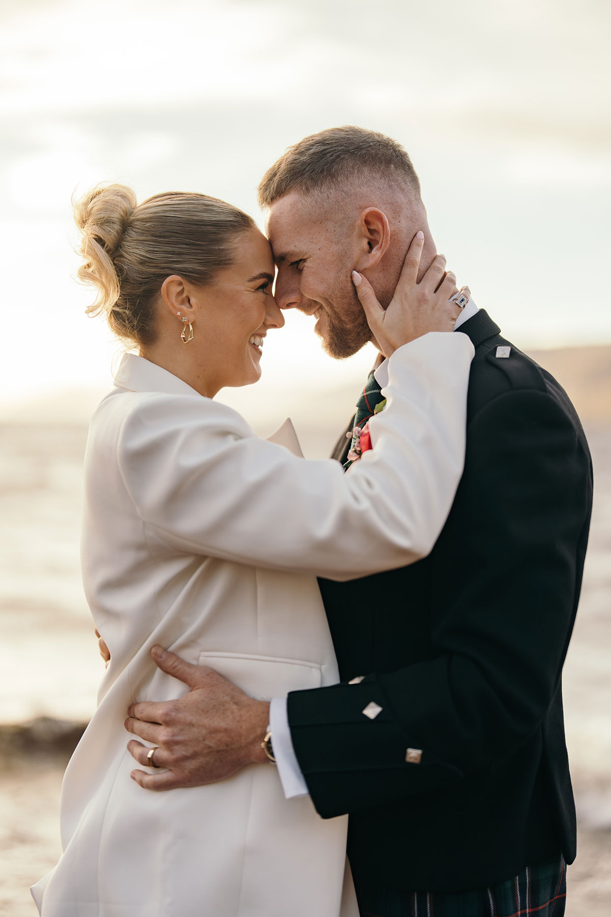 Bride in white suit and groom in kilt embrace during golden hour coastal wedding photos