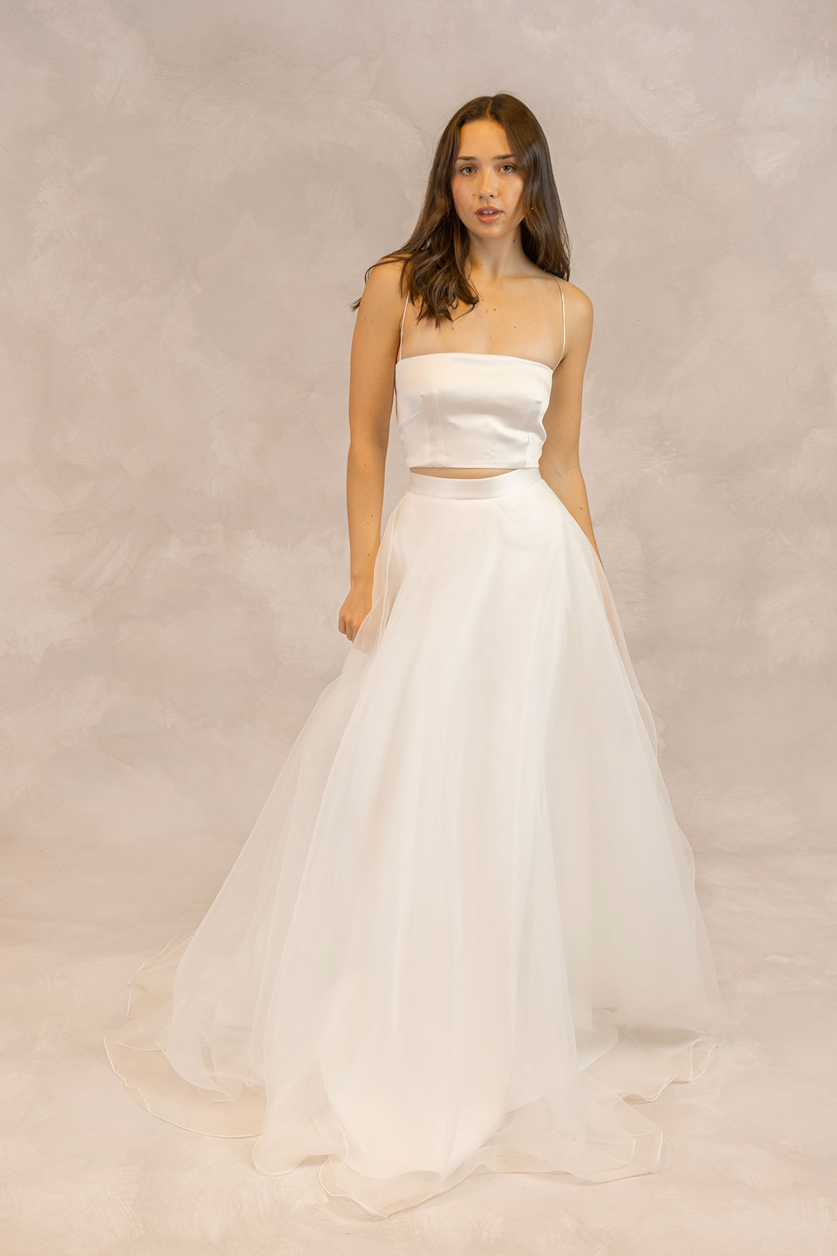 Model wearing a minimalist two-piece wedding look with a structured satin crop top, delicate spaghetti straps and a full tulle skirt, photographed against a soft neutral studio backdrop