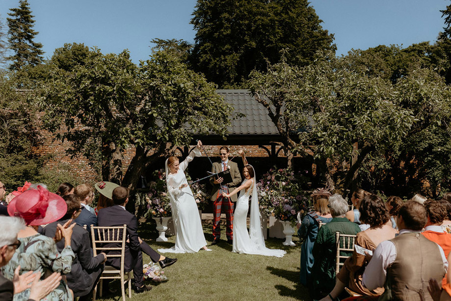 two brides performing a hand fasting with tartan fabric during their outdoor wedding at two brides walking through a confetti shower during outdoor wedding in the walled garden at the Byre at Inchyra