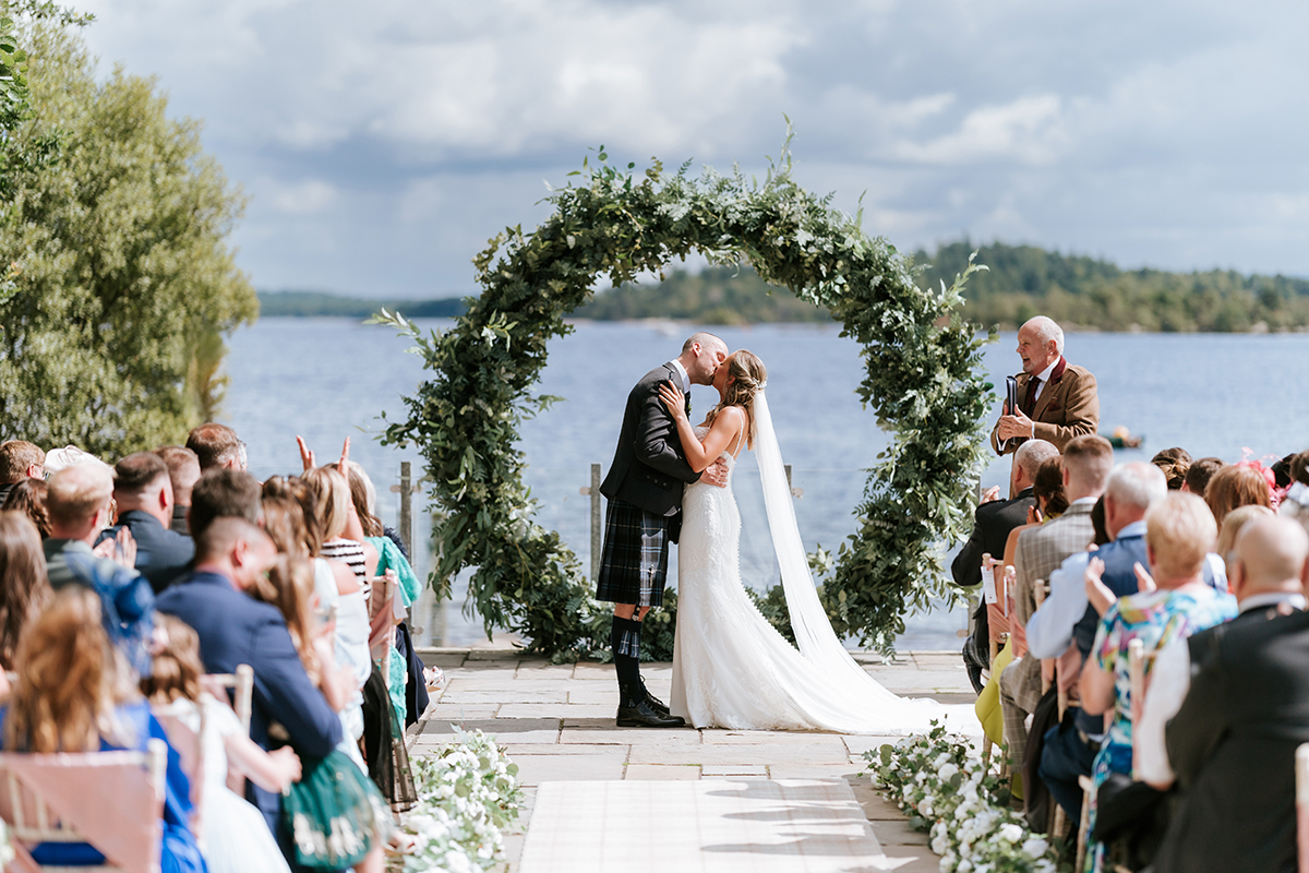 bride in trailing wedding dress with long veil and groom in dark blue kilt outfit kiss at top of aisle in front of circular greenery arch with view out across the water behind them
