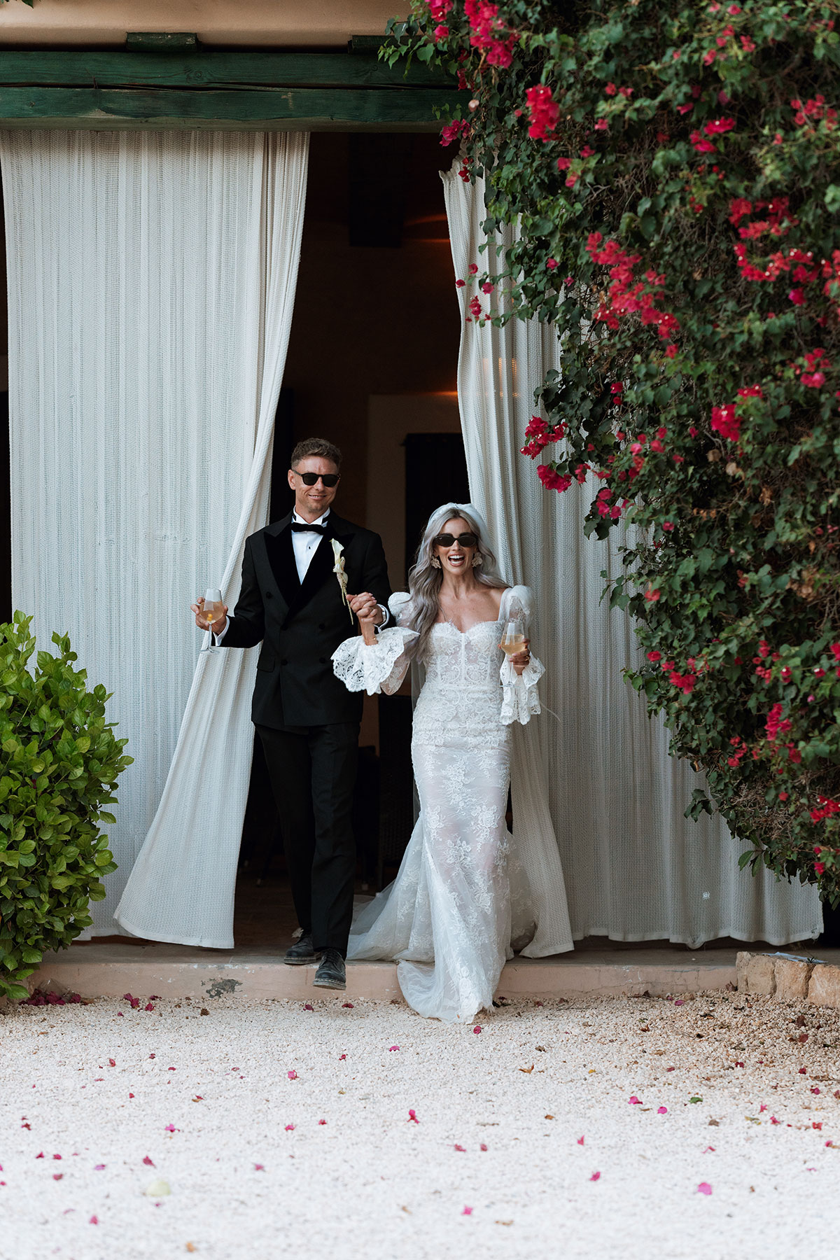 Bride and groom emerging through white curtains with champagne glasses at Can Rafal Ibiza wedding reception