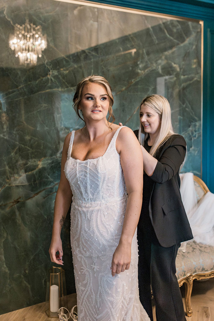 bride in beaded wedding dress has woman in black suit, buttoning up the back of her gown