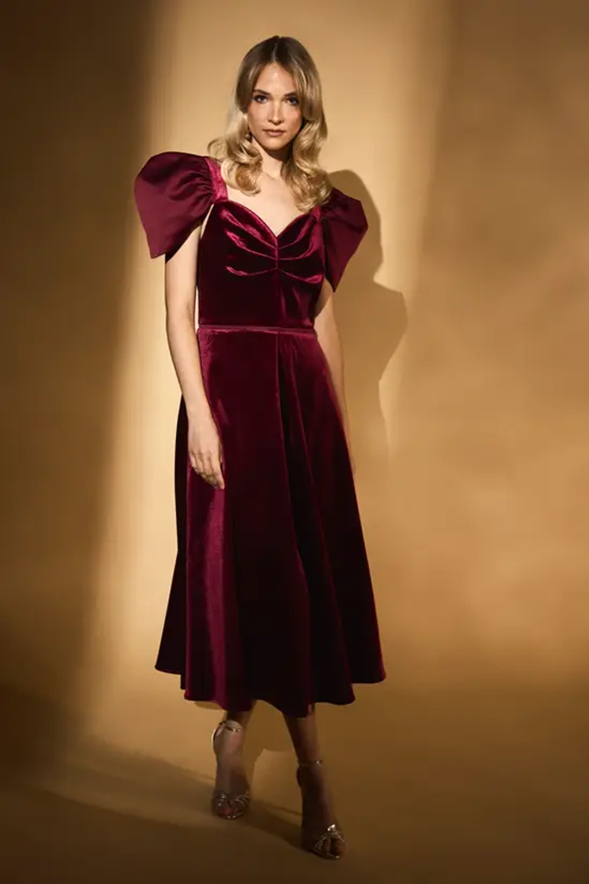 Woman wearing a burgundy velvet midi dress with a structured bodice, draped neckline and dramatic sleeves.