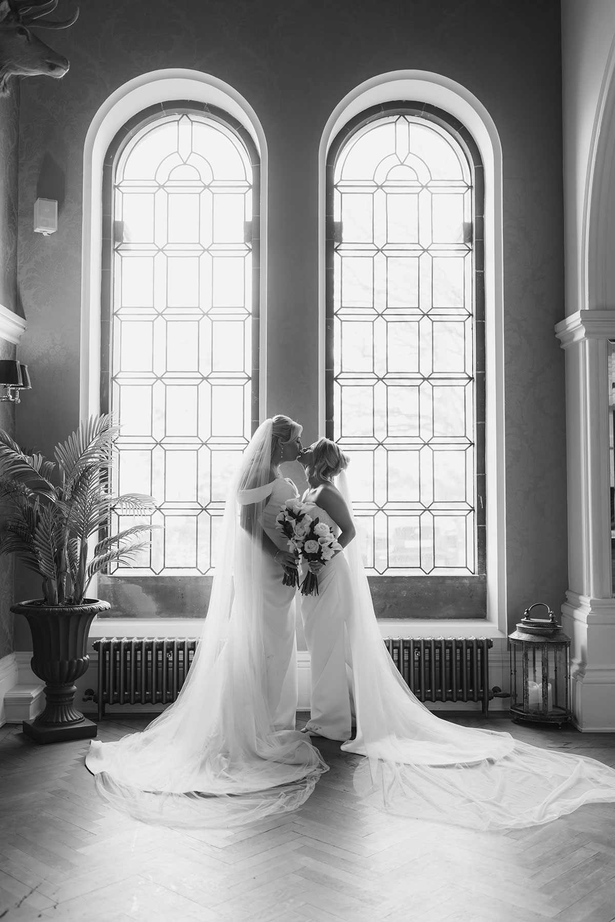 Black and white portrait of the newlyweds kissing in front of grand arched windows, their cathedral-length veils flowing behind them