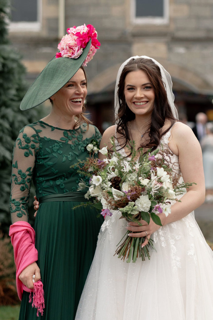 a smiling bride holding a large bouquet of green, purple and white flowers. She is standing next to a woman wearing a green dress with pink wrap and green hat with pink and red floral detail. They are standing in front of the Nethybridge Hotel