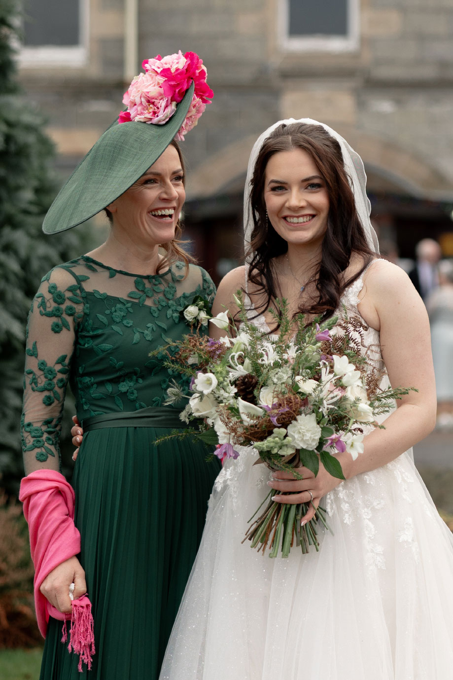a smiling bride holding a large bouquet of green, purple and white flowers. She is standing next to a woman wearing a green dress with pink wrap and green hat with pink and red floral detail. They are standing in front of the Nethybridge Hotel