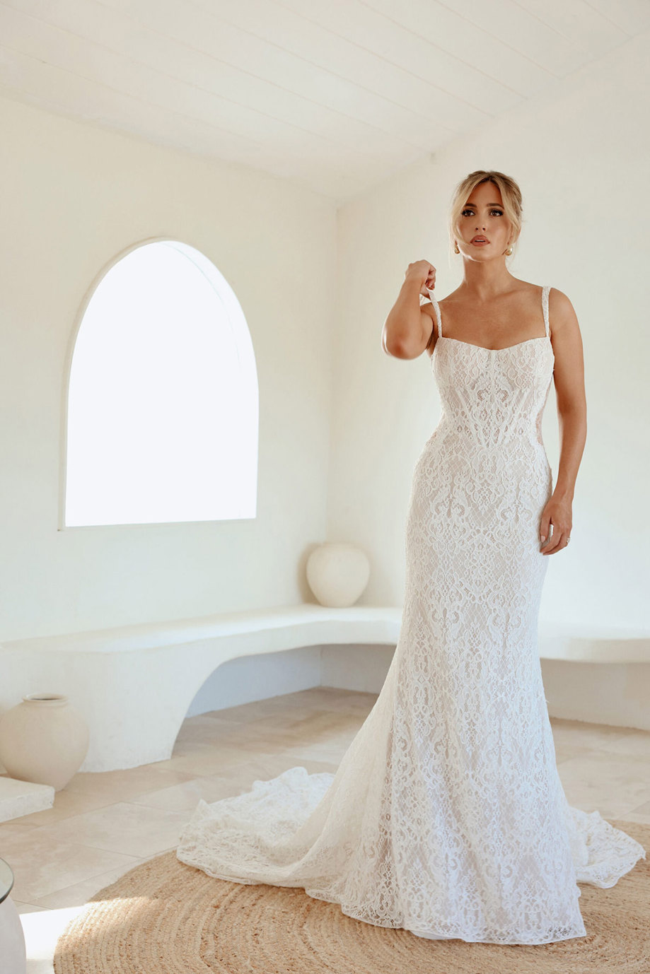 blonde woman in lace form fitting wedding dress with thin straps a train and her hair up with whispy hair along the side of her face