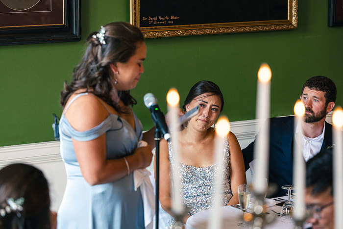 a seated bride and groom look emotional as a bridesmaid in a pale blue dress stands and looks at a phone while standing next to a microphone stand. There are white tapered candles flickering in the foreground