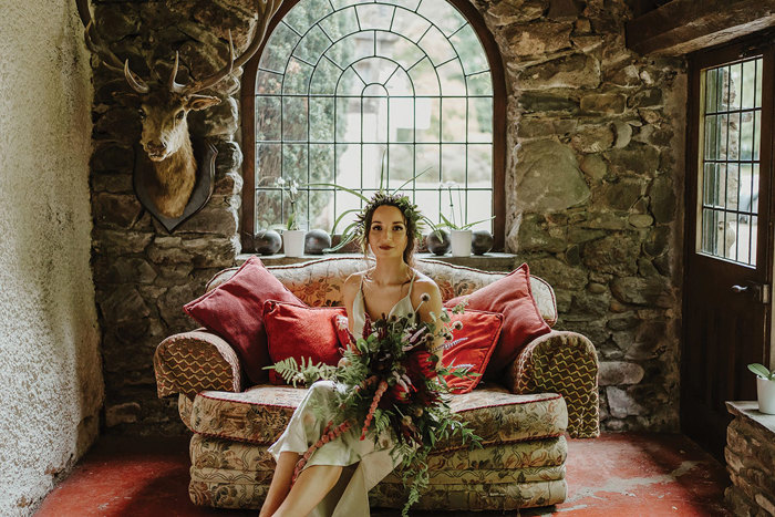 Bride sits on chair in rustic room holding pink and red bouquet