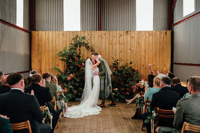 bride in flowy white wedding dress and long veil kisses groom in green and pale blue kilt at a flower arrangement altar while rows of people sit and clap