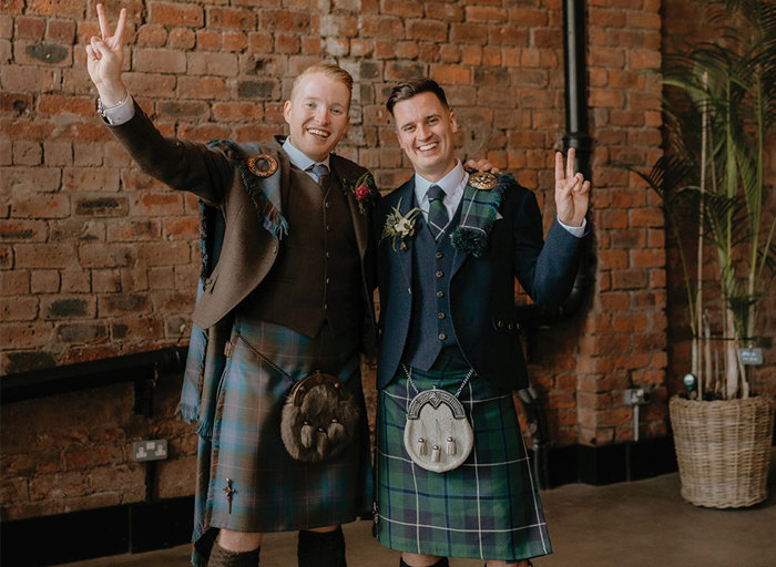 Two grooms with arms around each other wearing different coloured tartan kilts holding up the peace sign