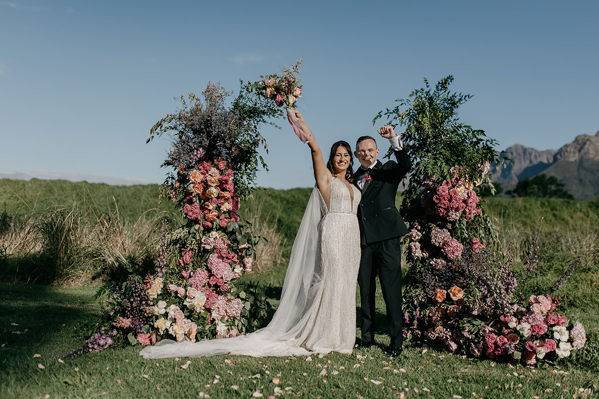 Newlyweds celebrating after outdoor ceremony beneath colourful floral installation at Boschendal Wine Estate wedding in South Africa