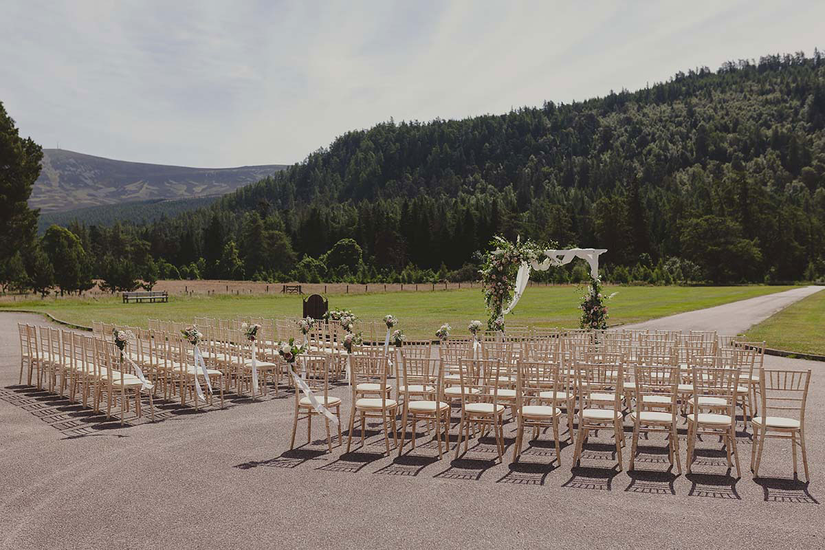 Outdoor wedding ceremony setup with chairs arranged in rows facing a floral arch, surrounded by green hills and forest