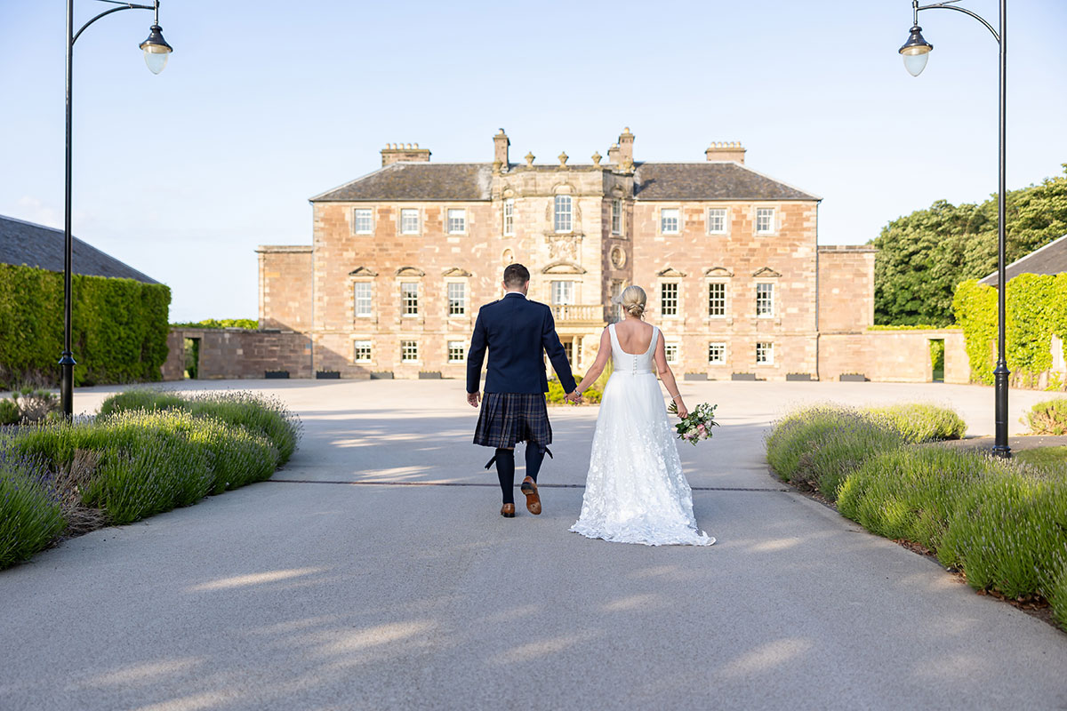 view from the back of bride and groom couple walking hand in up towards the front entrance of a large regal looking brick home