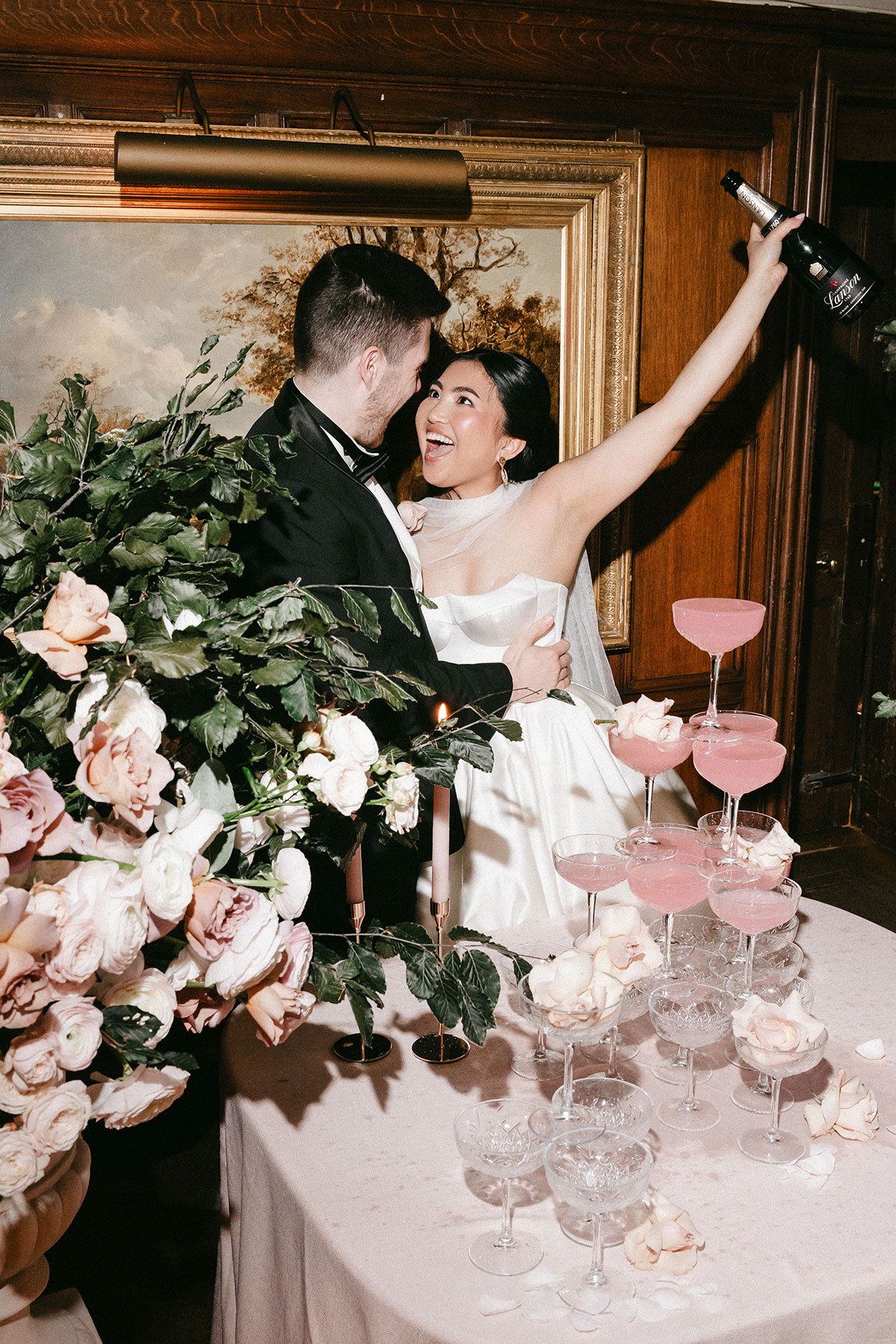 Newlyweds celebrating indoors beside a pink cocktail tower, the bride lifting a champagne bottle joyfully while surrounded by lush floral arrangements