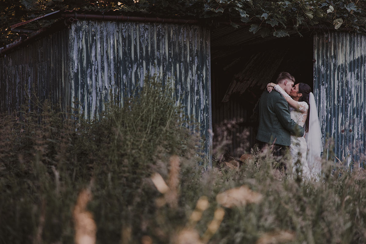 Bride and groom embracing outside a rustic corrugated metal shed surrounded by tall grass