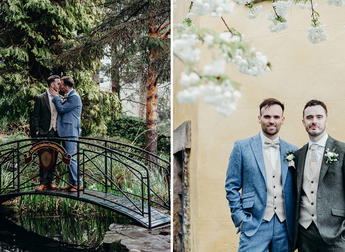 two grooms kissing on a small black arched bridge in a green garden on left. Two grooms standing against a yellow brick wall underneath some white cherry blossom on right