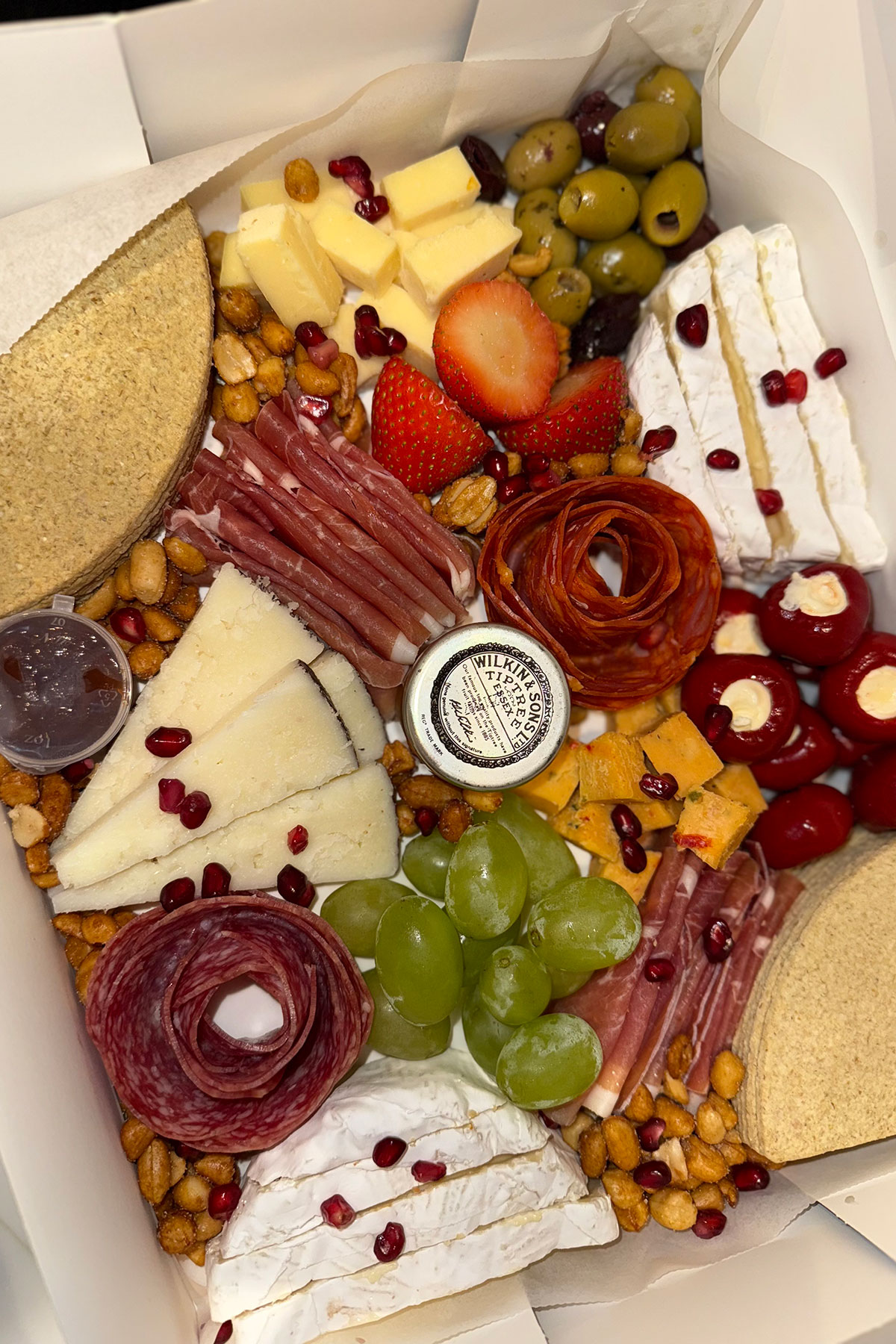 Close-up of an open grazing box featuring sliced cheeses, charcuterie, olives, strawberries, grapes, nuts and pomegranate seeds arranged neatly around a mini jar of chutney