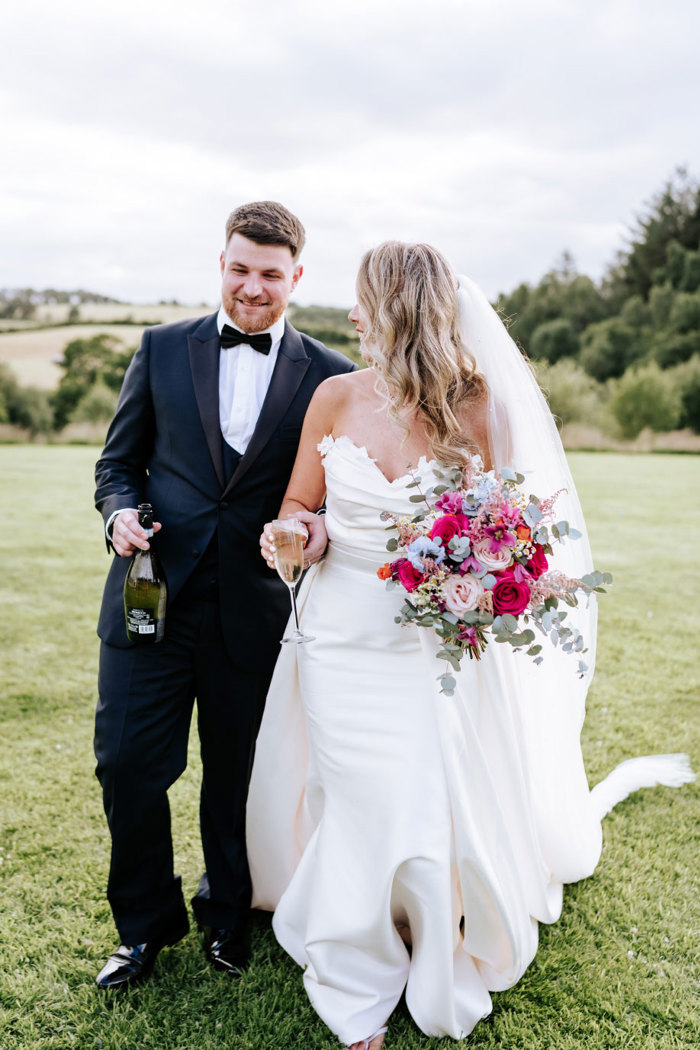 a bride and groom walking on grass. She is holding a champagne glass and he is holding a bottle of prosecco