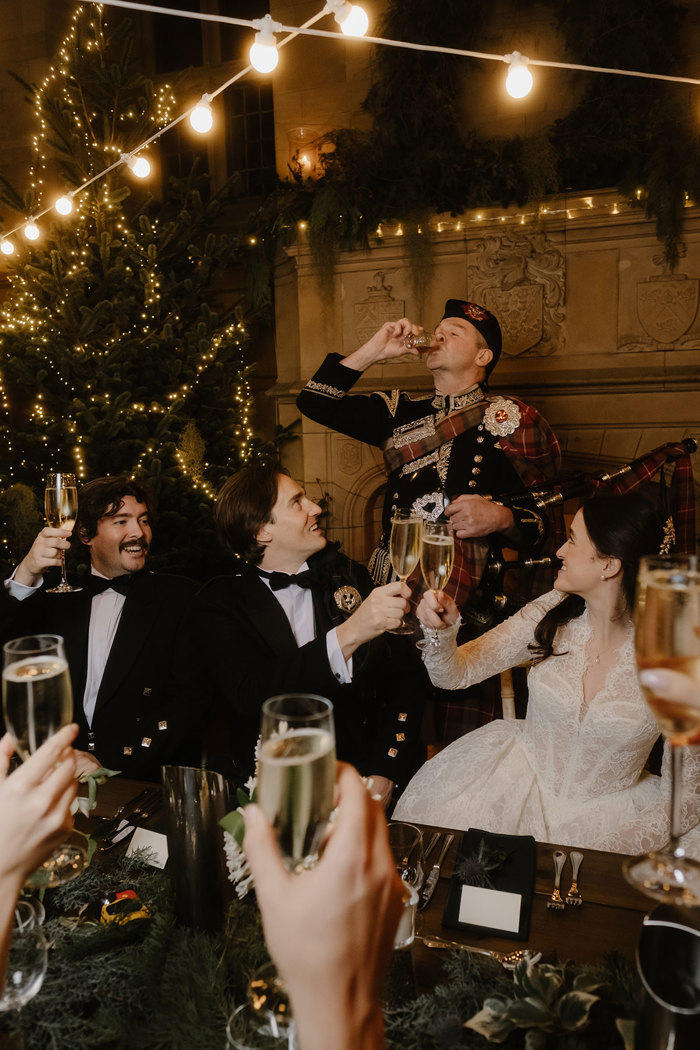 a bride and groom toasting champagne glasses in front of a man wearing Scottish formal attire.