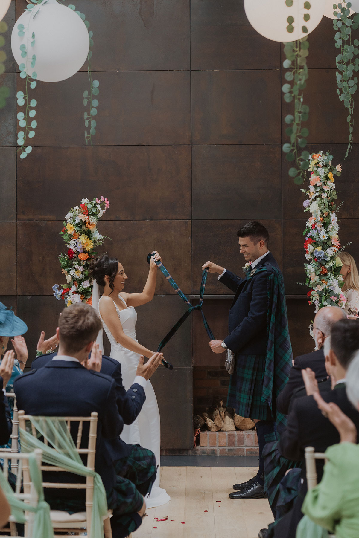 Bride and groom take part in traditional Scottish handfasting ceremony inside rustic barn-style venue.