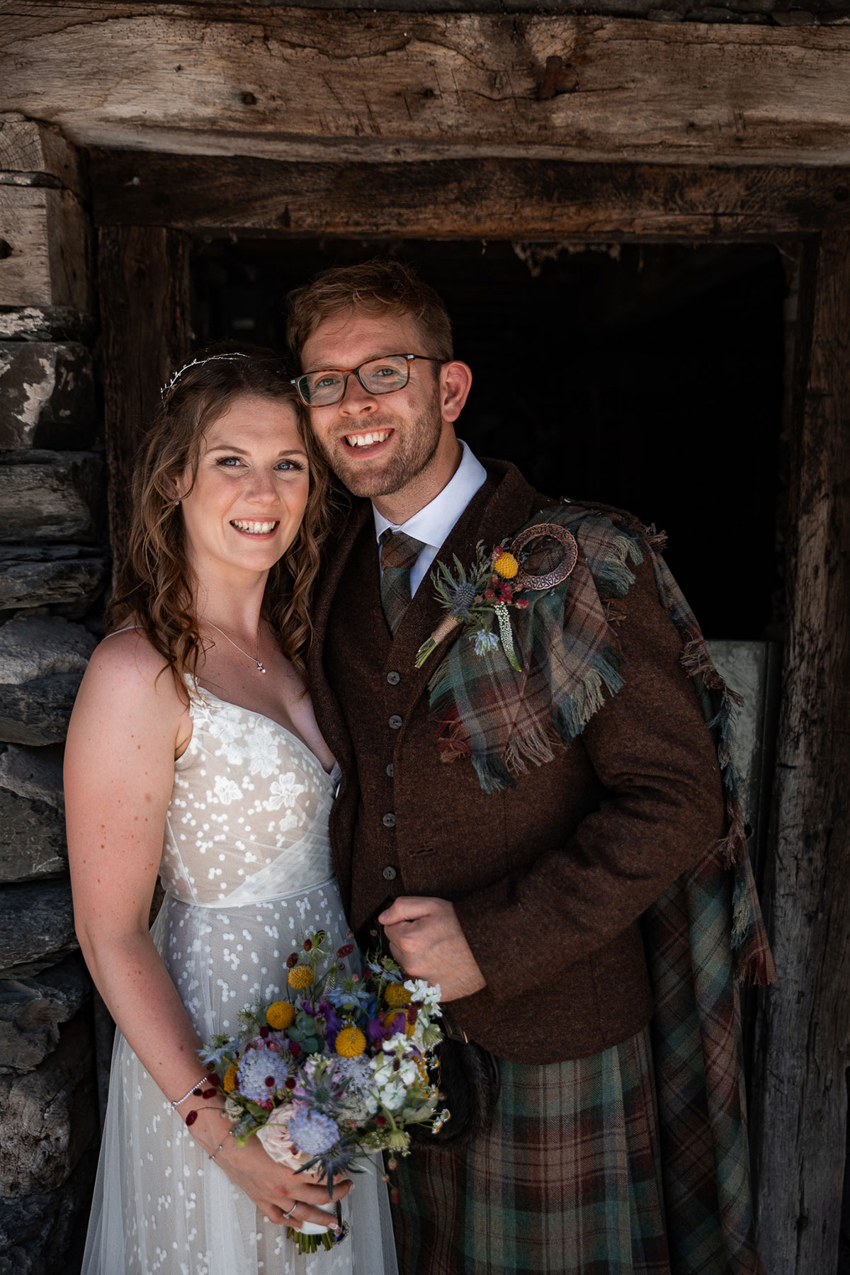 Portrait of bride and groom standing close together in a rustic stone doorway. The bride wears a lace wedding dress and holds a colourful bouquet, while the groom wears a brown tweed jacket with tartan kilt and fly plaid