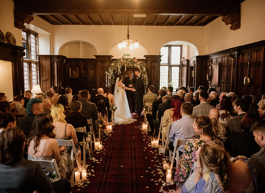 A wedding ceremony in a dark wooden panelled room looking up the aisle at the bride and groom holding hands
