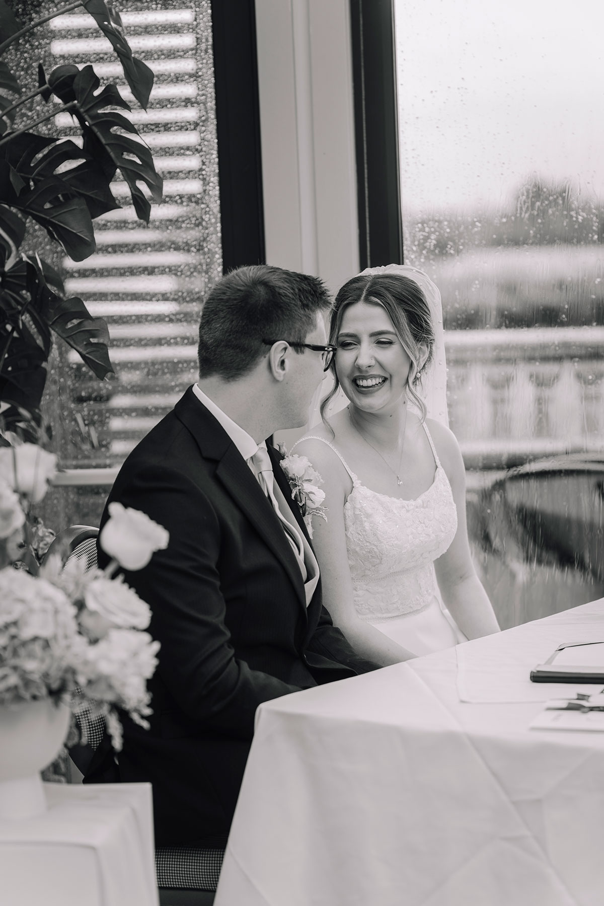 Bride and groom signing the marriage schedule together at a table by a rain-speckled window