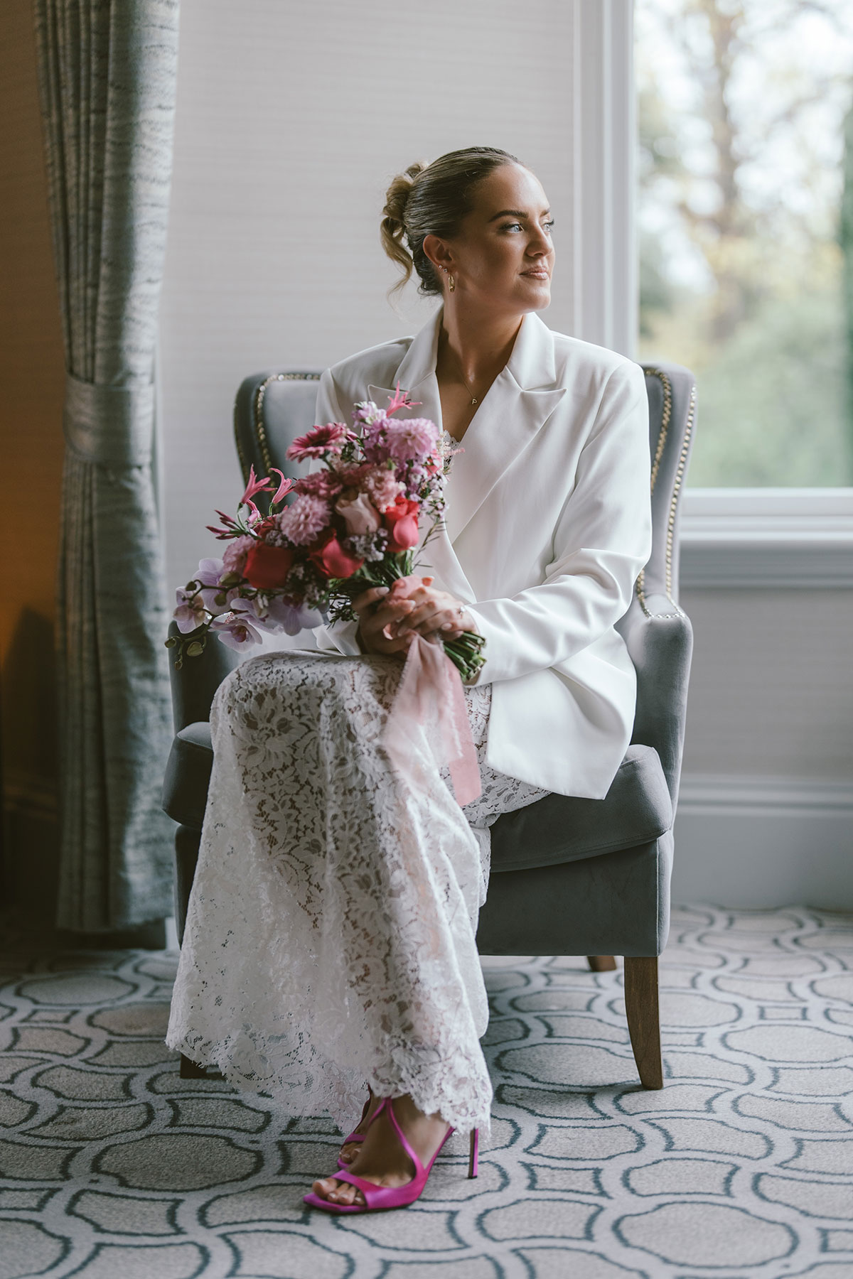 Bride in white suit holding pink bouquet seated by window in soft natural light
