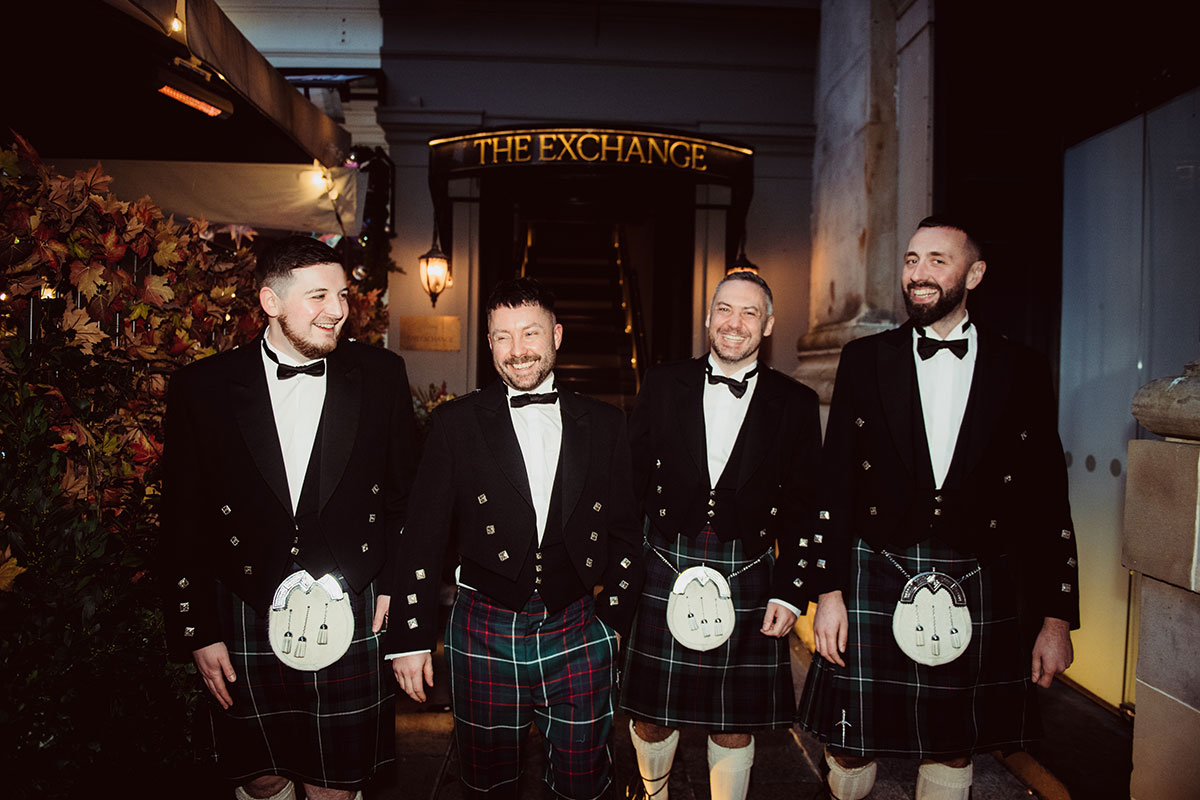 Group of men wearing formal jackets and kilts walking together outdoors at night