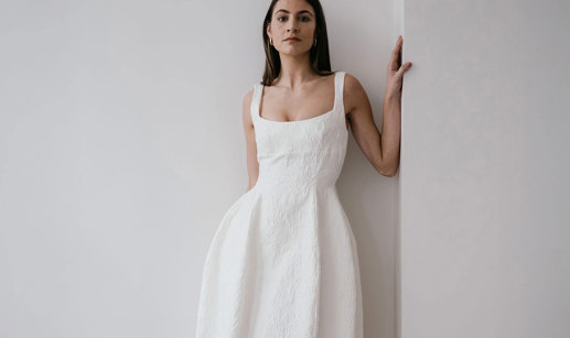 a dark haired model stands in the corner of two plain white walls, leaning to one side, her hand resting on the wall at shoulder height as she wears textured white wedding dress with a structured hourglass shape